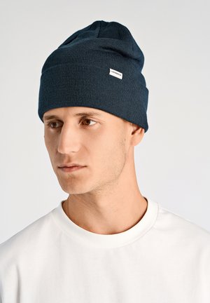 Dark blue knit beanie with a slightly slouchy design and a small white label. Paired with a plain white t-shirt. Smooth texture.