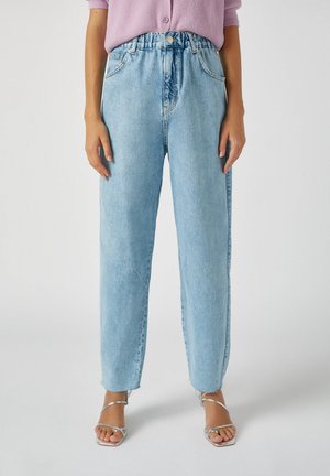 Kobieta nosząca jasnoniebieskie proste jeansy z wysokim stanem, elastycznym paskiem i guzikiem, w połączeniu z fioletową bluzką i srebrnymi sandałkami na obcasie z paskami.