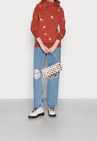 Rustrød hættetrøje med guldbelægninger i bladform, lyseblå slidte jeans og en hvid og brun ternet håndtaske. Sorte snøresko.