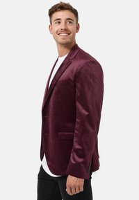 Blazer in velluto bordeaux con una texture morbida, chiusura a pulsante singolo e due tasche frontali. Abbinato a una t-shirt bianca.