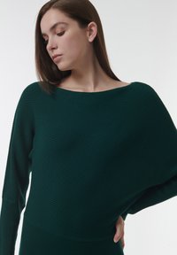Femme aux longs cheveux bruns portant un pull côtelé vert foncé à épaules dénudées, regardant vers le bas, une main posée sur la hanche.