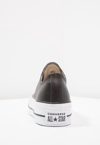 Černé kožené tenisky s bílou gumovou podrážkou, které mají nízký střih a viditelné logo "Converse All Star" na patě.