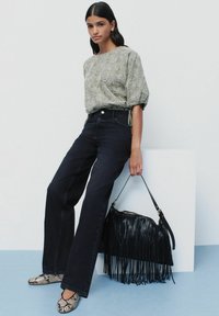 Blusa de patrón verde, jeans de pierna ancha oscuros y flats de serpiente. Bolso negro con flecos y correa para el hombro. Fondo liso.
