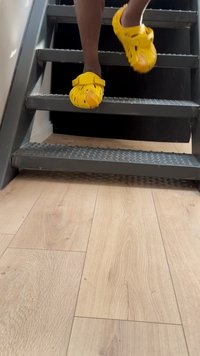 O persoană urcă niște scări metalice purtând încălțăminte galbenă strălucitoare, cu podeaua din lemn vizibilă dedesubt. Ambientul este bine luminat și interior.