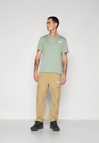 Vaaleanvihreä, lyhythihainen t-paita, jossa logo, yhdistettynä beigeihin cargohousuihin. Mustat vaelluskengät, seisomassa yksivärisen taustan edessä.