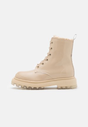 Bottine beige brillante avec semelle épaisse à crampons, devant à lacets avec six œillets, et doublure douce et pelucheuse visible au niveau du bord supérieur.