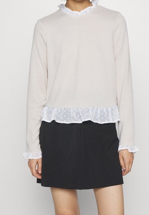 Haut beige clair à manches longues avec un col à volants blancs et un ourlet transparent à pois. Associé à une jupe sombre avec des poignets à volants.