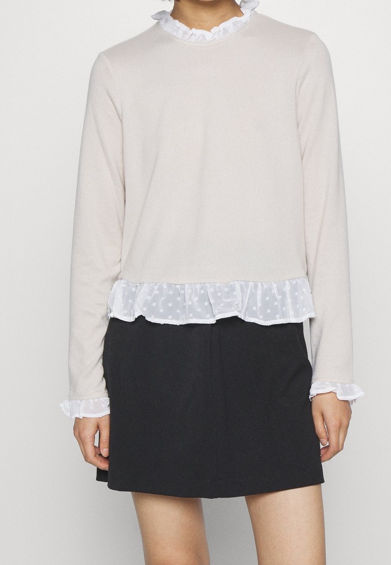 Helles beiges Langarmoberteil mit einem weißen Rüschensaum und transparentem Punkte-Saum. Kombiniert mit einem dunklen Rock, der Rüschenmanschetten hat.