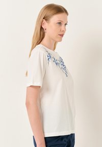 T-shirt bianco in cotone con maniche corte, caratterizzato da motivi floreali blu sulla parte anteriore. Il colletto è rotondo e la vestibilità è comoda.