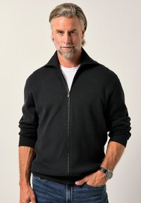 Černý svetřík na zip s vysokým límcem, žebrovanými manžetami a lemem. Nositelný přes bílou košili, kombinovaný s modrými džíny. Má hladkou texturu.