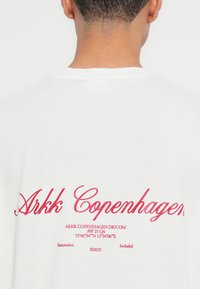 Valge T-särk ümarate kaelustega, mille seljal on punane tekst, sealhulgas "Arkk Copenhagen" ja täiendavad brändi detailid all.