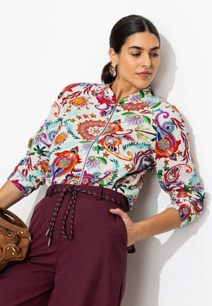 Femme aux cheveux foncés en queue de cheval, portant un chemisier zippé fleuri coloré et un pantalon taille haute bordeaux, tenant un sac à main en daim marron.