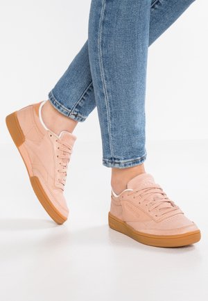 Osoba nosící světle růžové nízké tenisky s gumovou podrážkou a modré džíny, stojící na bílém podlaze.