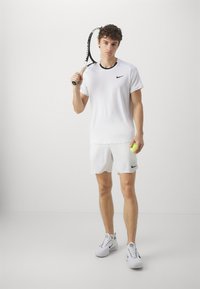 Hvit trenings t-skjorte med svart krage og Nike-logo, sammen med hvite shorts med svart logo. Holder en tenniskølle og en ball.