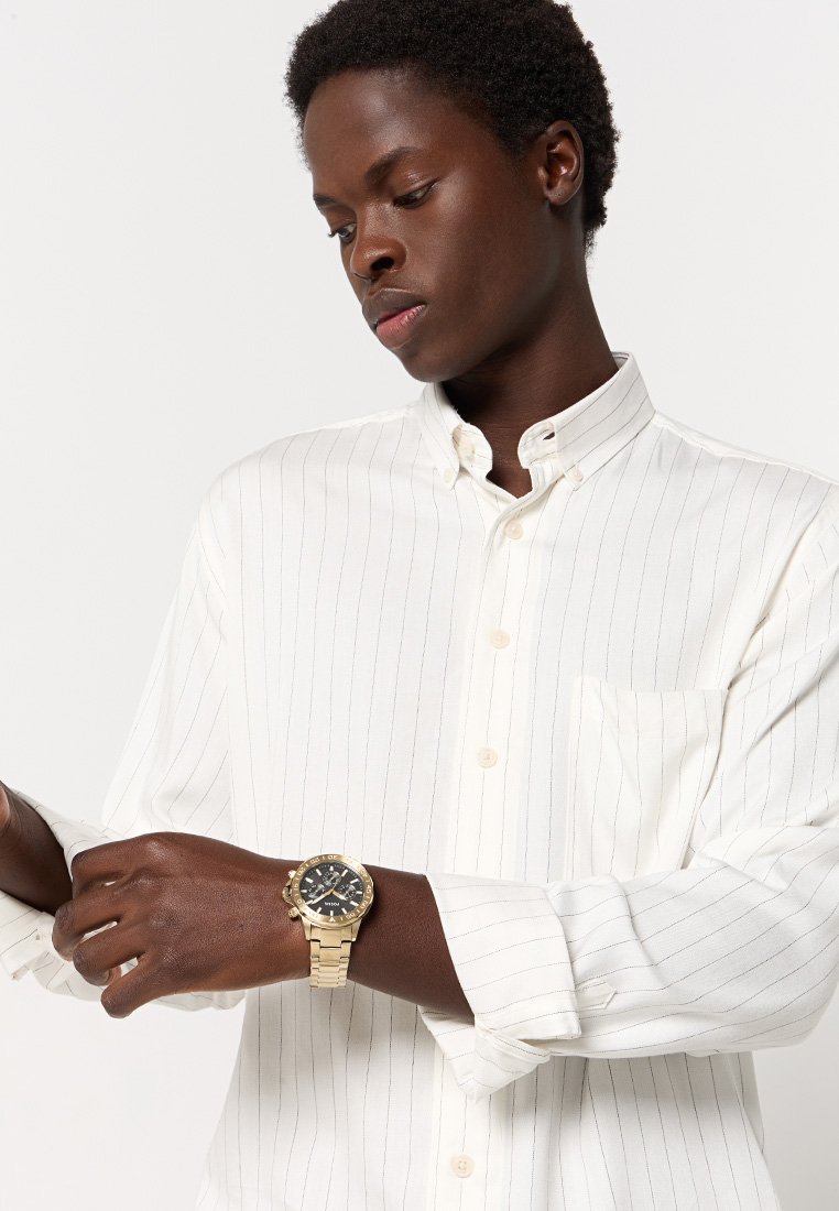 Giovane che sistemava la manica di una camicia bianca a righe, indossando un orologio d'oro con quadrante nero sul polso sinistro, sfondo neutro.