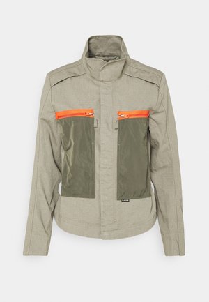 Veste marron clair avec col montant, deux grandes poches avant olive et des fermetures éclair orange vif sur chaque poche.