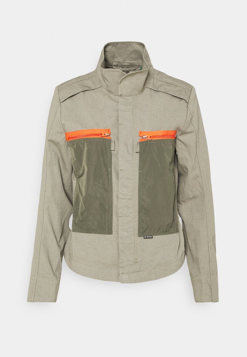 Veste marron clair avec col montant, deux grandes poches avant olive et des fermetures éclair orange vif sur chaque poche.