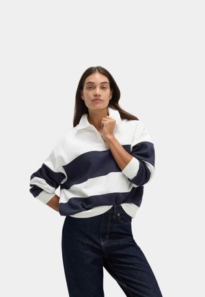 Frau trägt ein navy-weiß gestreiftes Poloshirt mit langem Arm und Kragen, dunkelblaue Jeans, posiert mit einer Hand nahe am Kinn und der anderen an der Hüfte.