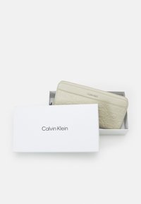 Calvin Klein Geldbörse in hellbeige mit geprägtem Logo-Muster. Verpackt in einer weißen Box, die den Markennamen in Schwarz zeigt.