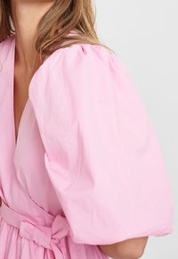 Rosa klänning med puffärmar, djup V-ringning och matchande bälte i samma tyg. Tillverkad av ett lätt material med en slät textur.