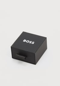 Boîte rectangulaire noire avec un onglet, portant un logo blanc "BOSS" sur la surface supérieure. Texture lisse, design minimaliste.