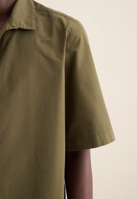 Camicia verde oliva a maniche corte realizzata in un tessuto liscio, con colletto classico e orlo dritto e pulito, con dettagli di cucitura minimi.