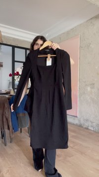 Personne tenant une robe noire à manches longues sur un cintre dans une pièce moderne avec un sol en bois et des murs texturés.