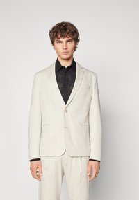 Lys beige blazer med et subtilt rutemønster, enkelt knappelukking, og notched krager, parret med en svart mønstrete skjorte.