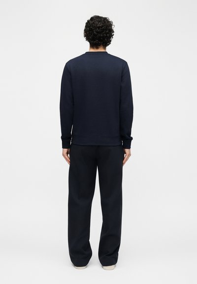 Άνδρας με σγουρά μαλλιά που φοράει ένα μπλε σκούρο φούτερ και μαύρο παντελόνι με φαρδιά μπατζάκια, στέκεται στραμμένος σε έναν κενό λευκό τοίχο.