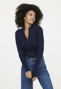 Marineblauer, gerippter Strickpullover mit hohem Kragen und halbem Reißverschluss. Lange Ärmel reichen über die Handgelenke und sind mit blauen, hoch-taillierten Jeans kombiniert.