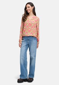 Femme souriante, portant un haut à manches longues rose à motifs, un jean large bleu clair et des chaussures à plateforme noires, se tenant sur un fond blanc.