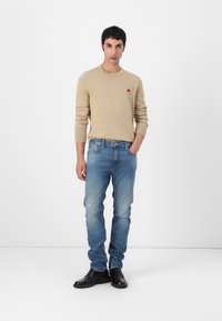 Beiger Pullover mit kleinem rotem Logo, kombiniert mit hellblauen verwaschenen Jeans und schwarzen Dressschuhen. Outfits werden vor einem weißen Hintergrund präsentiert.