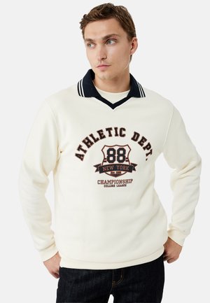 Joven con sudadera blanca con cuello negro y ribete a rayas, con el texto "Athletic Dept. 88 New York Championship".