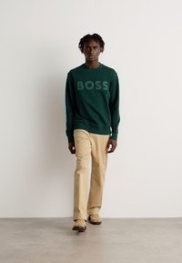 Felpa verde con logo "BOSS", abbinata a pantaloni beige; il modello cammina su uno sfondo neutro, caratterizzato da tessuti testurizzati.