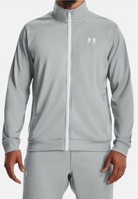 Ljusgrå träningsjacka med dragkedja och hög krage, vit dragkedja och broderad logotyp på vänster bröst. Tyget har en slät textur.