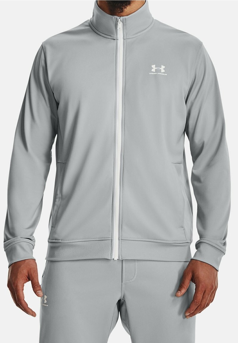 Ljusgrå träningsjacka med dragkedja och hög krage, vit dragkedja och broderad logotyp på vänster bröst. Tyget har en slät textur.
