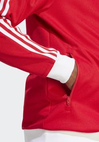 Red athletic jacket featuring white striped accents on the sleeves, with a zippered pocket near the side and ribbed white cuffs.