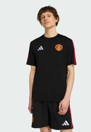 Ung mand iført sort Manchester United sportstøj med røde striber og Adidas-logoer, stående mod en ensfarvet lys baggrund.