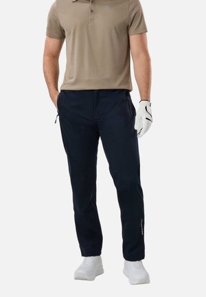 Mężczyzna ubrany w beżową koszulkę polo, granatowe spodnie z kieszeniami na zamek, białą rękawiczkę golfową i białe tenisówki, stojący z jedną ręką w kieszeni.