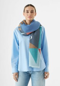 Écharpe géométrique multicolore avec des nuances de bleu, de turquoise et de marron en tissu doux, drapée autour du cou, complétée par une chemise à boutons bleu clair.