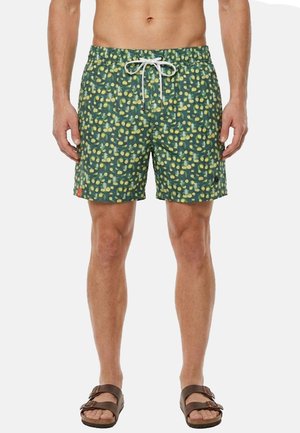 Uomo che indossa pantaloncini da nuoto verdi con motivo a puntini gialli e coulisse bianca, abbinati a sandali marroni con doppio cinturino.