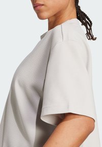 Camiseta de manga corta de color gris claro, texturizada, con un corte holgado, que presenta un escote redondo y detalles cosidos en las costuras de los hombros.