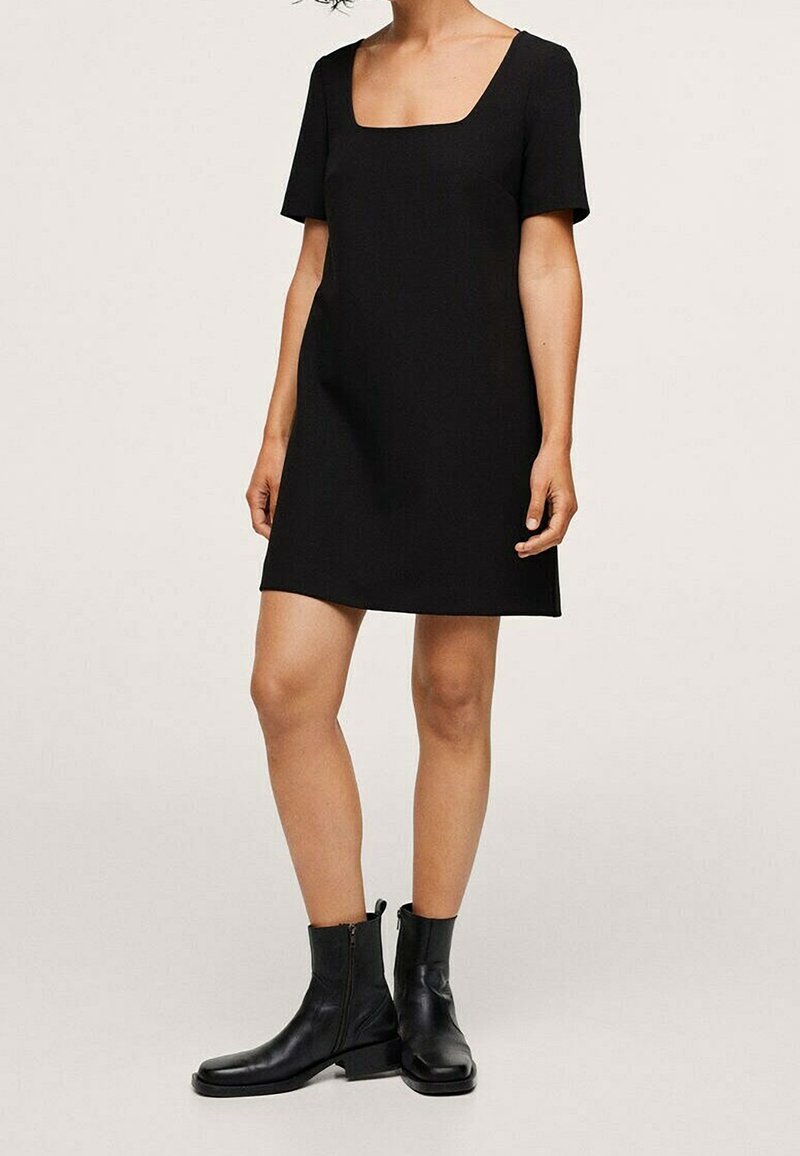 Femme portant une robe noire à manches courtes avec un décolleté carré, associée à des bottines noires sur un fond uni.