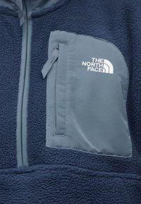 Tmavomodrá mikina z flísu s svetlomodrým zipsom na ebonovom vrecku na hrudi a výšivkou loga "The North Face" v bielej farbe.