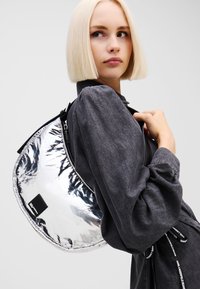 Metallic sølv halvmåneveske med en blank finish, svart glidelås og justerbar stropp. Modellen har på seg en mørk grå bluse.