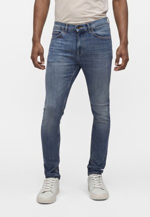 Homme portant un jean slim bleu et des baskets blanches, debout devant un fond clair uni.