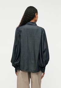 Tumšs denima krekls ar apkakles izgriezumu, pūkainām piedurknēm un brīvu piegriezumu. Gluda tekstūra ar smalku spīdumu un sānu slēdzenēm apakšmalā.