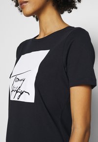 Camiseta de algodón negra con un gráfico rectangular blanco que presenta un diseño de firma en negro. Mangas cortas y cuello redondeado.