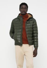 Olive green puffer jacket with a zip front and hood. Features a smooth texture, quilted design, and ribbed cuffs. Worn over a rust-colored turtleneck.