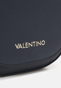 Granatowa skórzana torebka o zaokrąglonym kształcie, z metalicznym złotym logo "VALENTINO" oraz teksturowaną powierzchnią z subtelnymi detalami przeszyć.